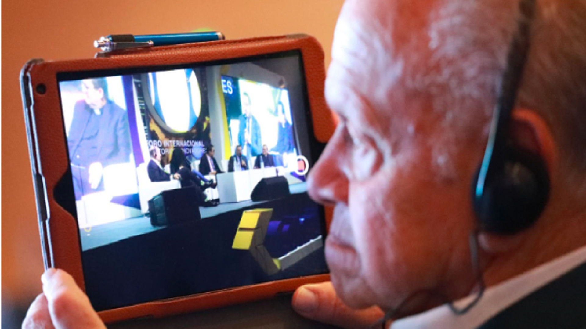 Adulto mayor con audífonos sostiene una tableta en la que se reproduce un video.