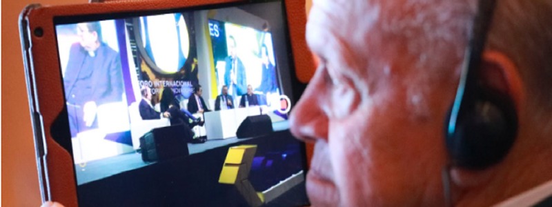 Adulto mayor con audífonos sostiene una tableta en la que se reproduce un video.