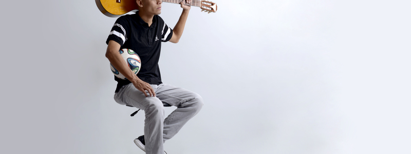 Jose Quiceno sostiene un balón bajo su brazo derecho y su guitarra detrás de su cabeza tomada por el mástil con su mano izquierda
