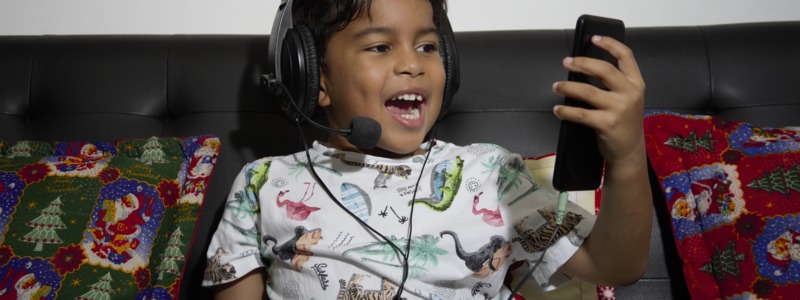 Fotografía: Niño sentado en un sofá usa audífonos de tipo diadema y habla frente a un celular
