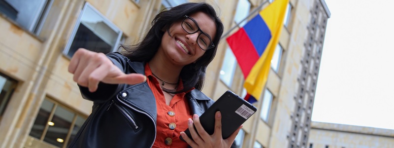 Fotografía: Mujer con celular en la mano y bandera de Colombia en la parte de atrás.