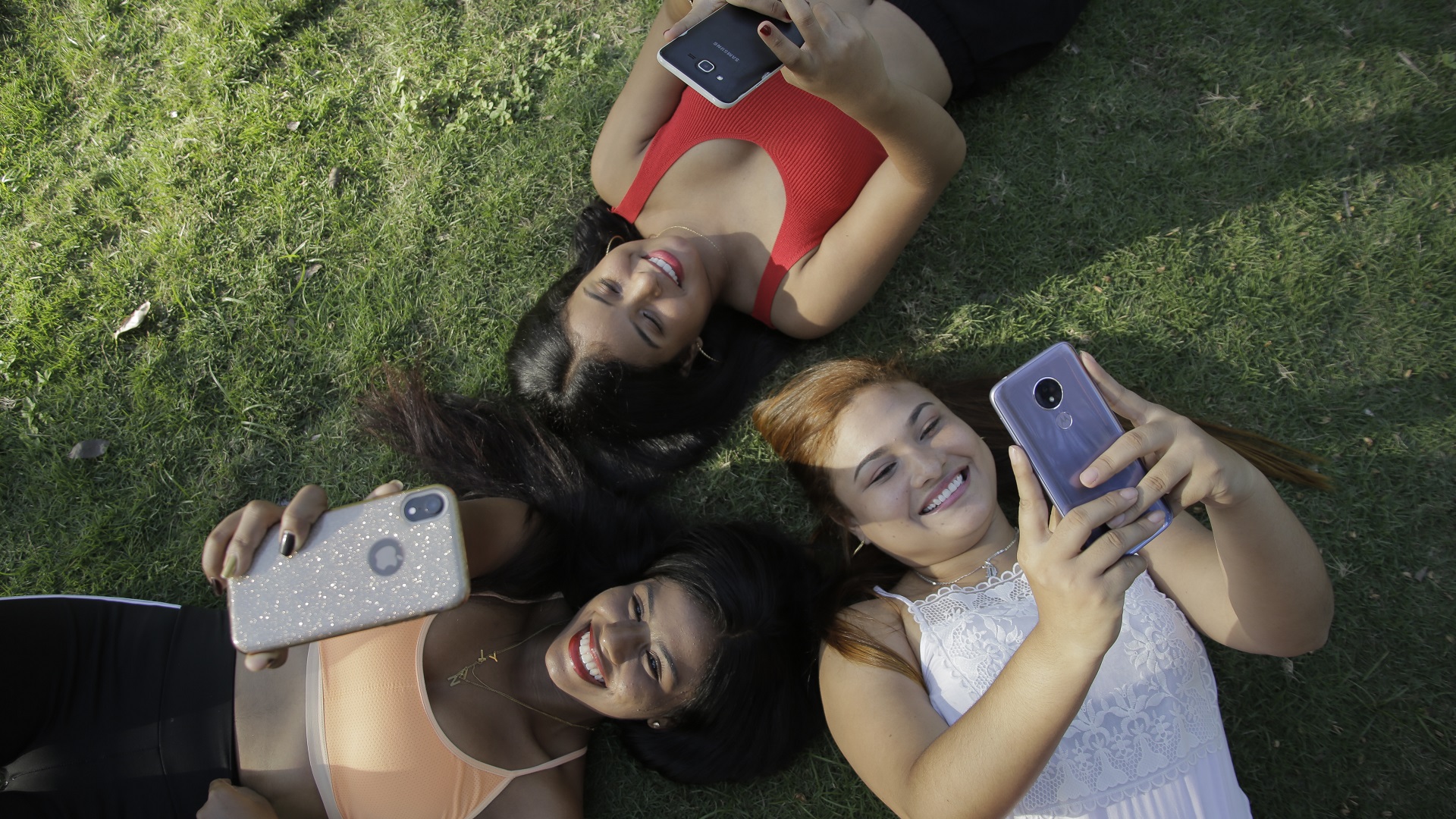 Tres mujeres jóvenes acostadas en el césped alzan sus manos y juegan con sus dispositivos móviles